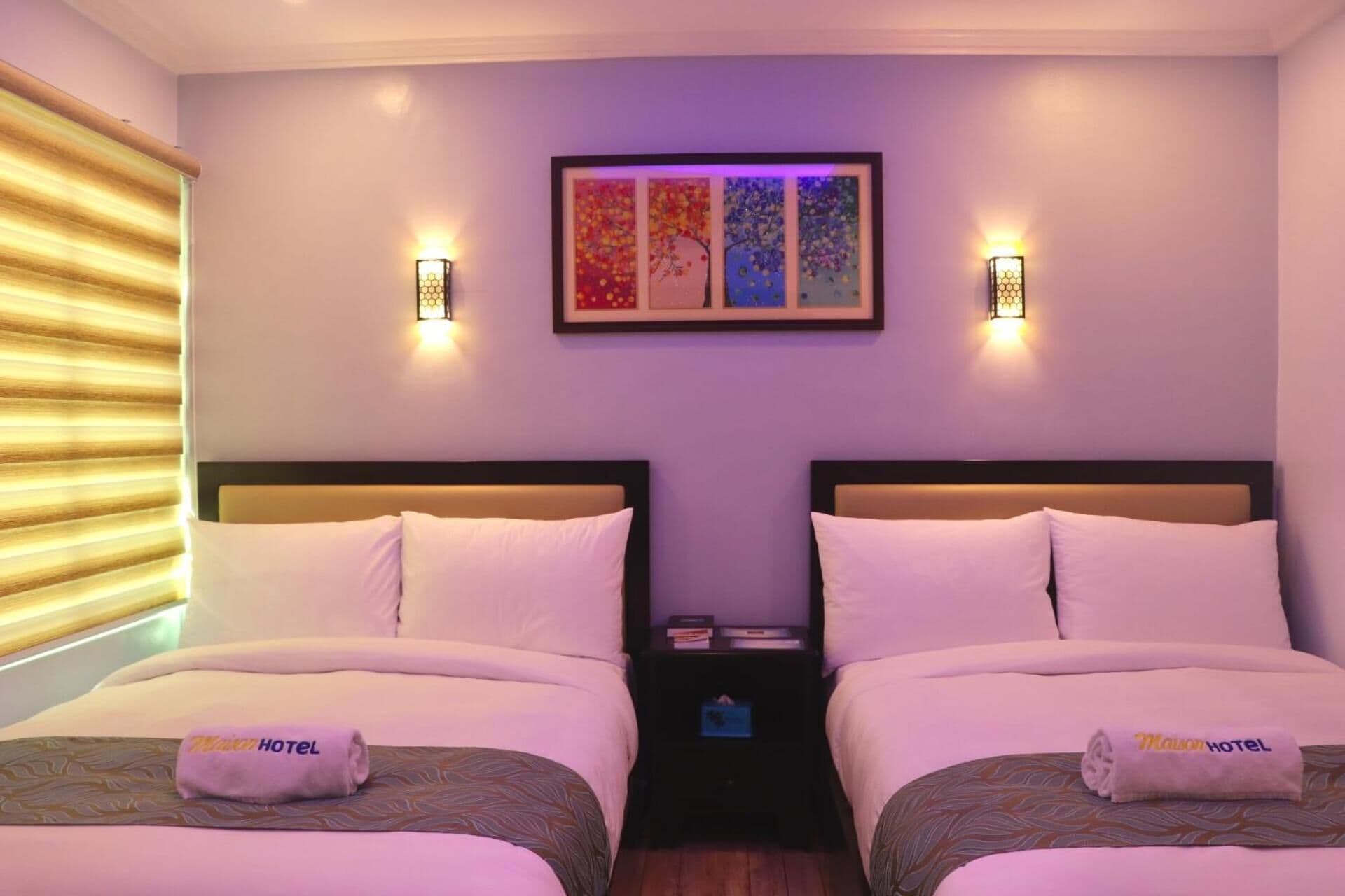 A hotel room with two beds, each with a pillow and a towel with the hotel name. There are wall-mounted lights and a framed artwork above the beds.