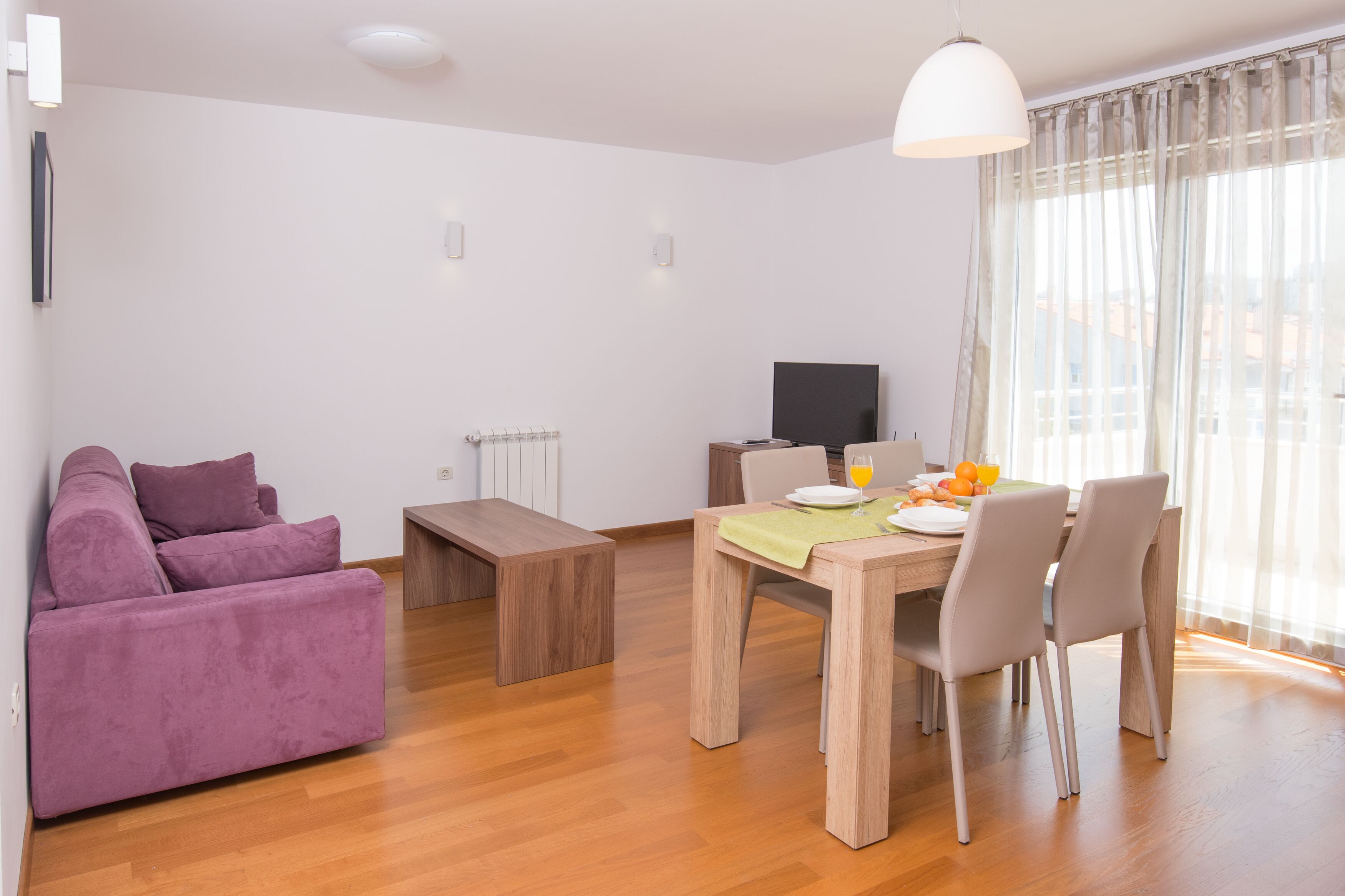 A modern living room with a purple sofa, a wooden dining table with chairs, a bench, a TV, and a window with sheer curtains.