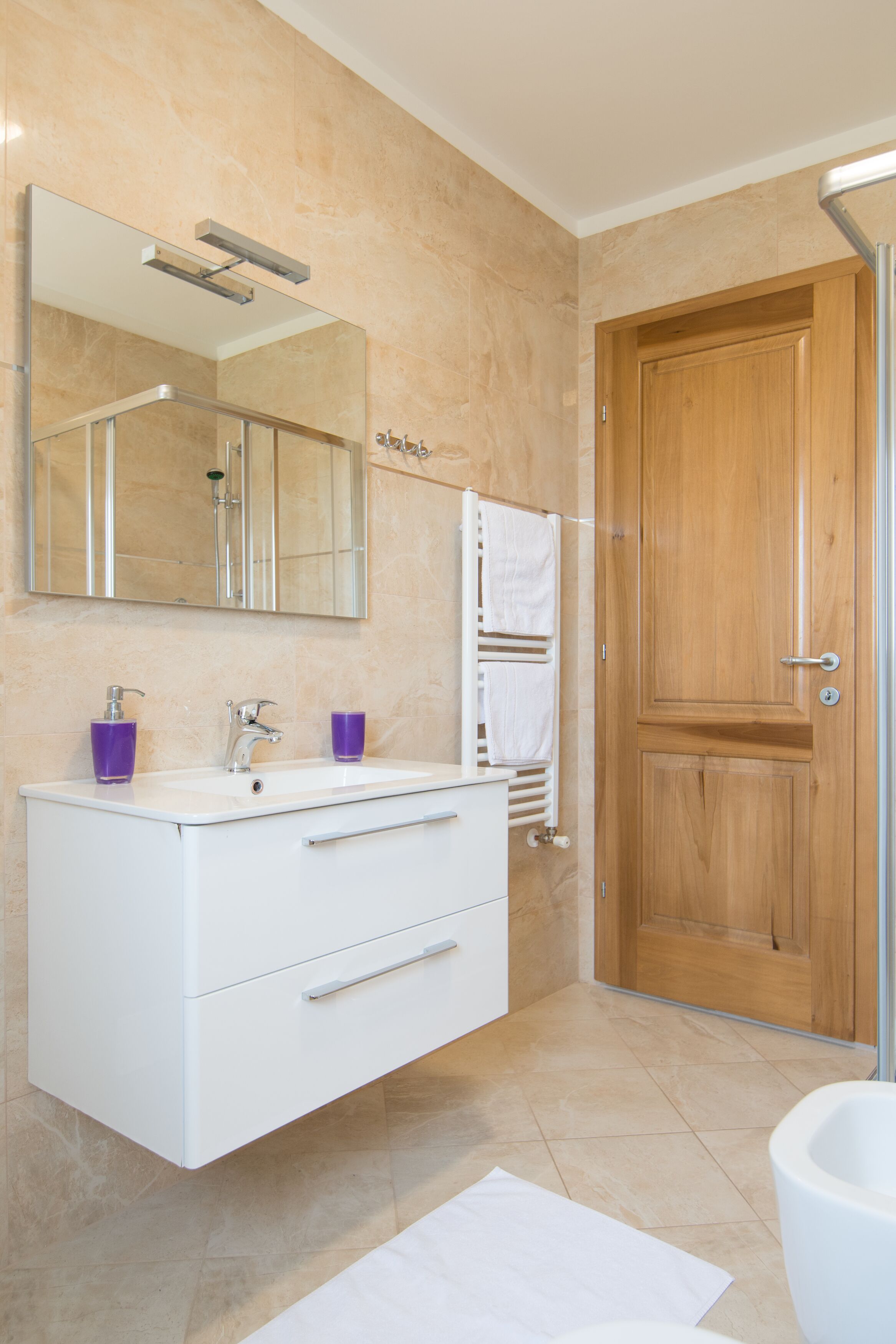 A modern bathroom with a white vanity, a large mirror, a wooden door, and a white bathtub.