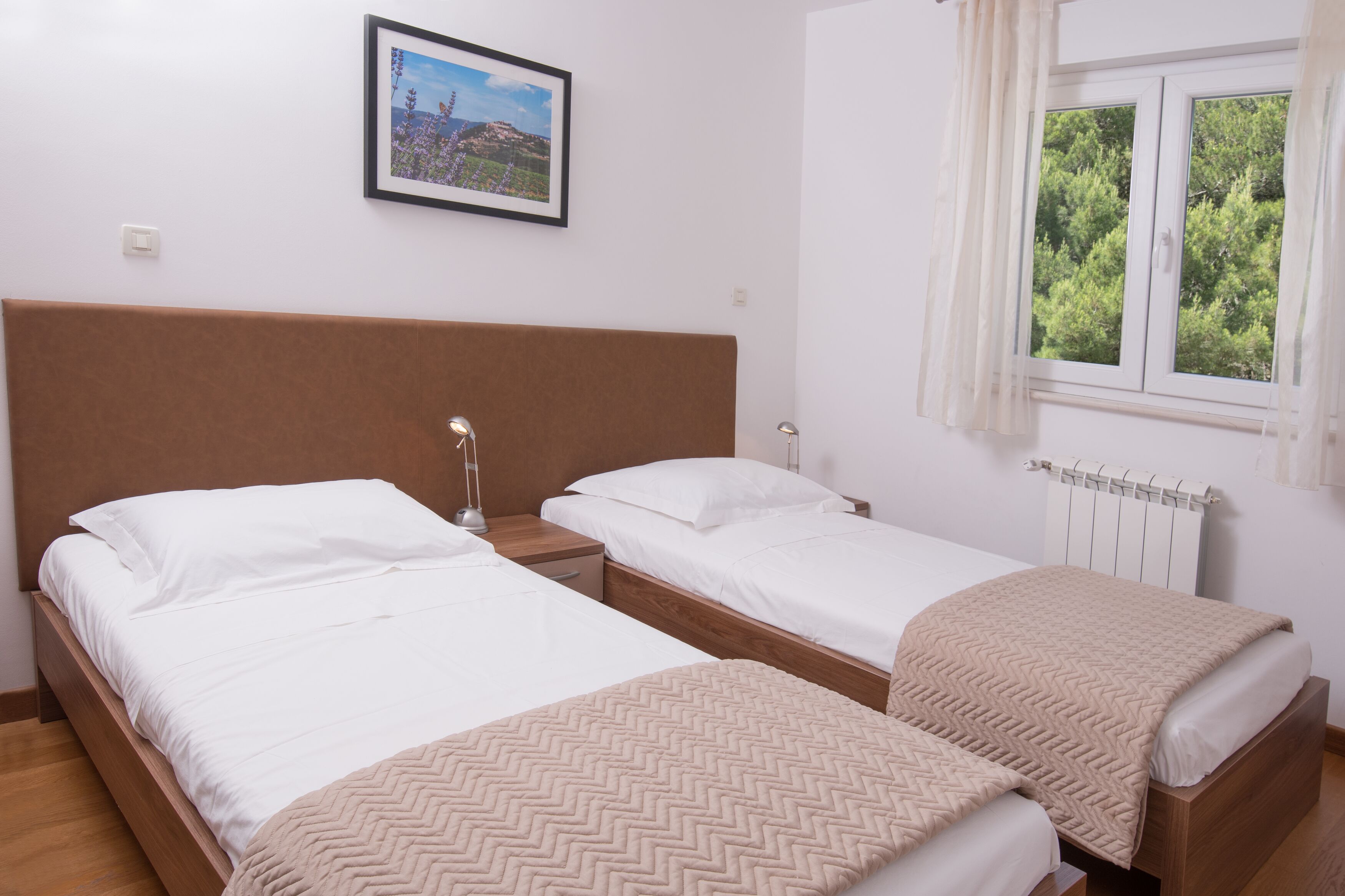 Una habitación con dos camas individuales, una mesita de noche con lámpara, un cuadro enmarcado en la pared y una ventana con vistas a árboles.