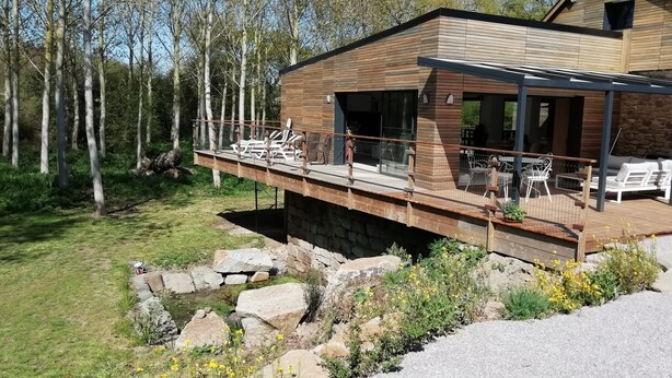 Una casa moderna con terraza de madera que se extiende sobre terreno rocoso, con mobiliario exterior y un muro de piedra.