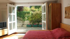 A bedroom with a bed, a television on a stand, and a view of a garden through an open door. - Apartment 2 in the beautiful Gutenberg / Ostallgäu (Gutenberg)