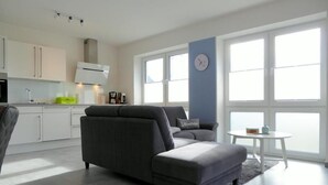 A modern living room with a grey sofa, a small round table, and a kitchen area with white cabinets and a blue accent wall.