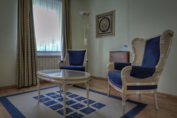 A room with two wicker chairs with blue cushions, a small glass table, a radiator, a wall clock, and a window with curtains. - Hostellerie le Prieuré de Conques (Florenville)