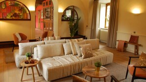 A cozy living room with a large beige sofa, a wooden coffee table, and a small side table. There is a potted plant on the side table and a chair with a cushion. - Hostellerie le Prieuré de Conques (Florenville)