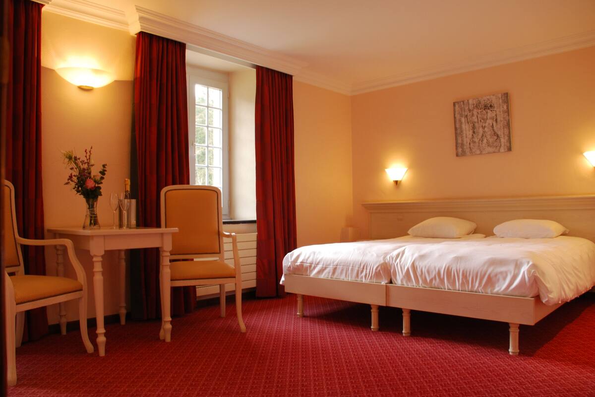 Habitación de hotel con cama, escritorio con silla, un florero con flores y ventana con cortinas.