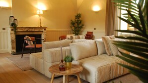 A living room with a beige sectional sofa, a round wooden coffee table with a candle and a plant, a fireplace with a lamp, and a potted plant. - Hostellerie le Prieuré de Conques (Florenville)