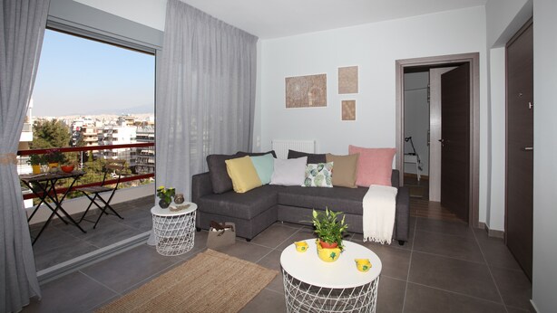 A modern living room with a grey sectional sofa, a round coffee table with decorative items, a balcony with a dining area, and a view of the cityscape. - Edem Beach Apartment with Mountain view by Athenian Homes (Paleo Faliro)