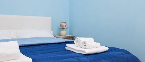 A neatly made bed with blue bedding and white pillows, a small bedside table with a lamp, and a blue wall.