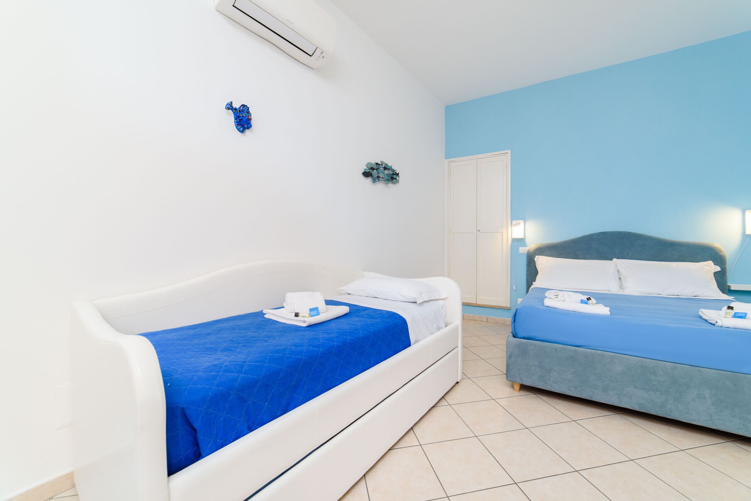 A hotel room with two beds, one with a blue bedspread and the other with a white bedspread. There is a white wall with a small decorative item and an air conditioning unit.