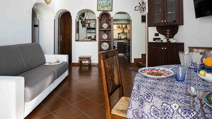 A living room with a grey sofa, a dining table with a blue patterned tablecloth, and a kitchen visible through an archway. - Namily House (Positano)