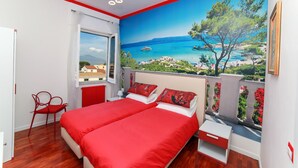 Un dormitorio con dos camas, una silla roja, una mesita de noche y una ventana con vista al paisaje costero.