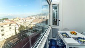 Un balcón con vista a la ciudad, una mesa con frutero y una ventana que da a edificios y montañas.