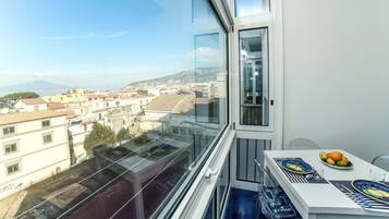 A balcony with a city view, a table with a fruit bowl, and a window with a view of buildings and mountains.
