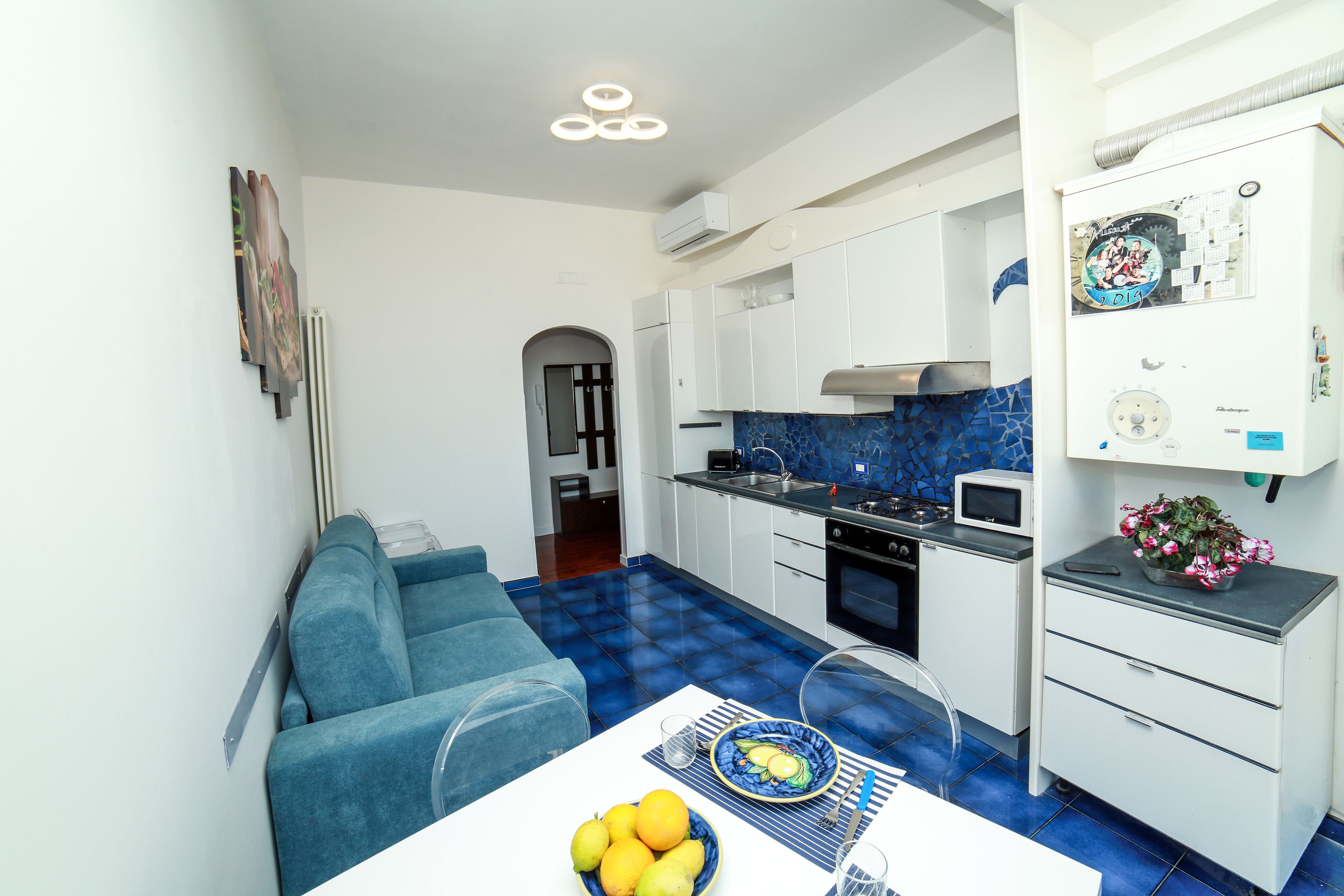 A modern kitchen with white cabinets, a blue tiled floor, and a dining area with a blue sofa and a table set with fruit.