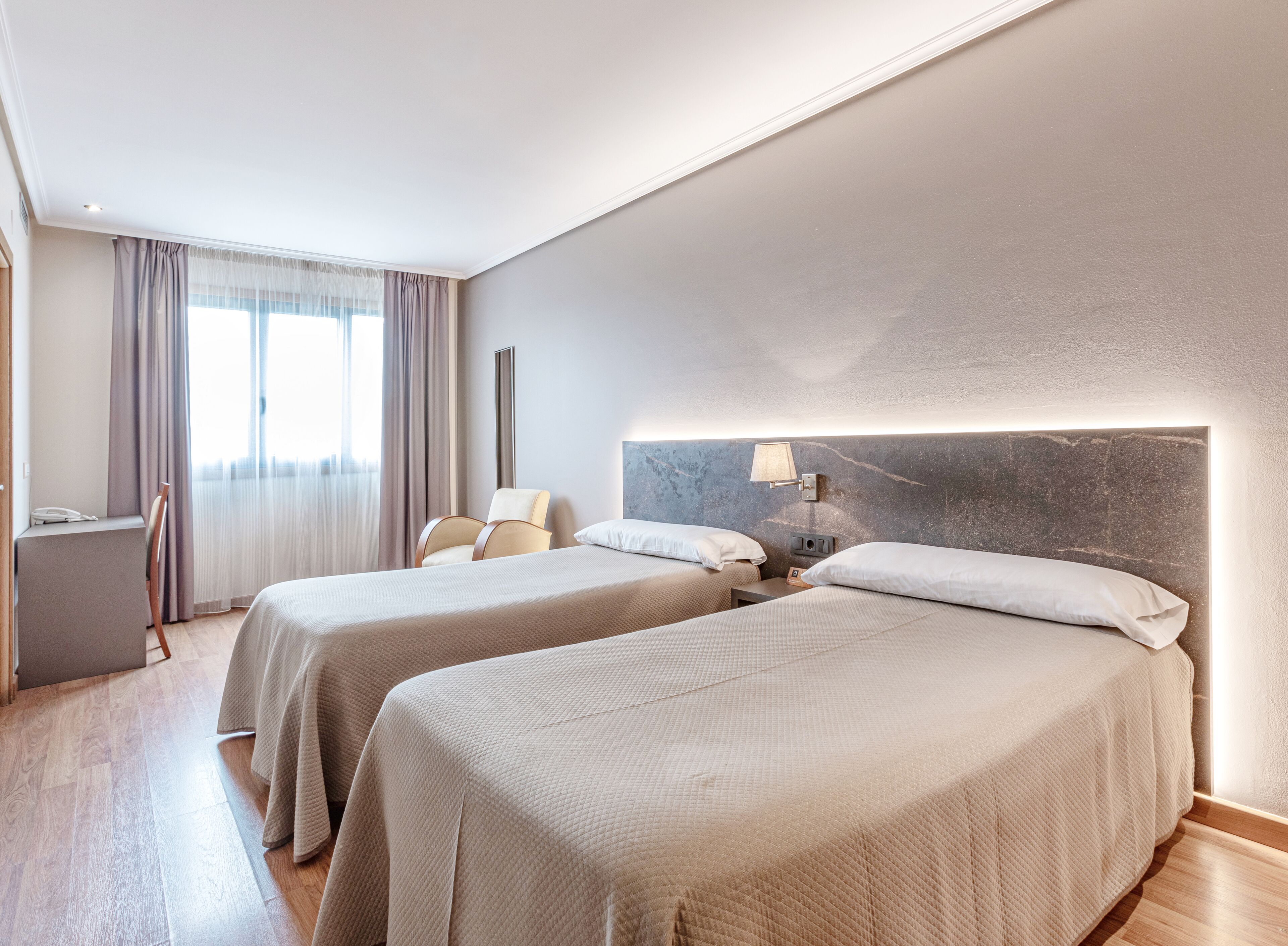 Quarto de hotel com duas camas, uma escrivaninha, uma cadeira e uma janela grande com cortinas.