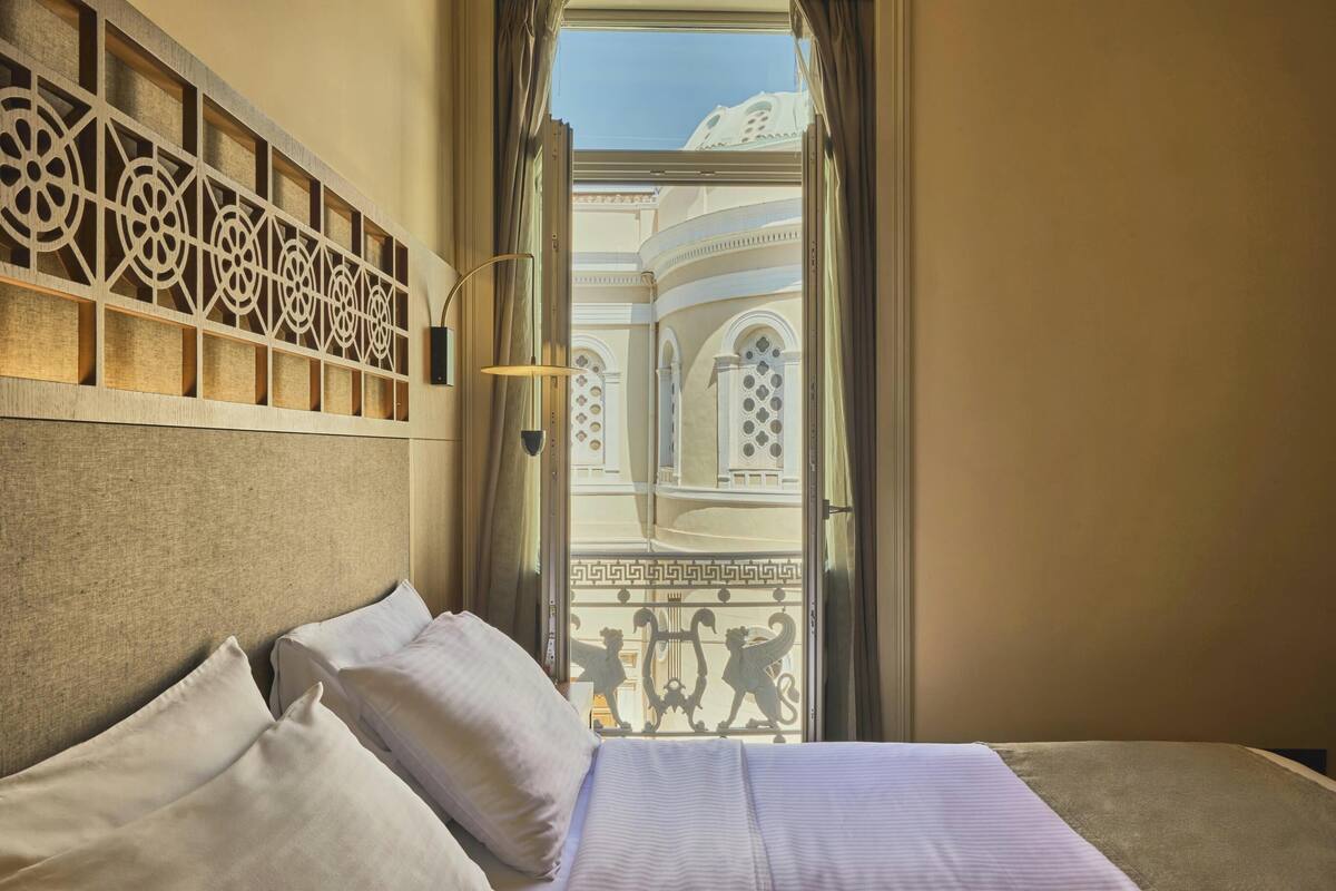 A hotel room with a bed, a decorative headboard, a window with a view of a building, and a balcony with a railing.