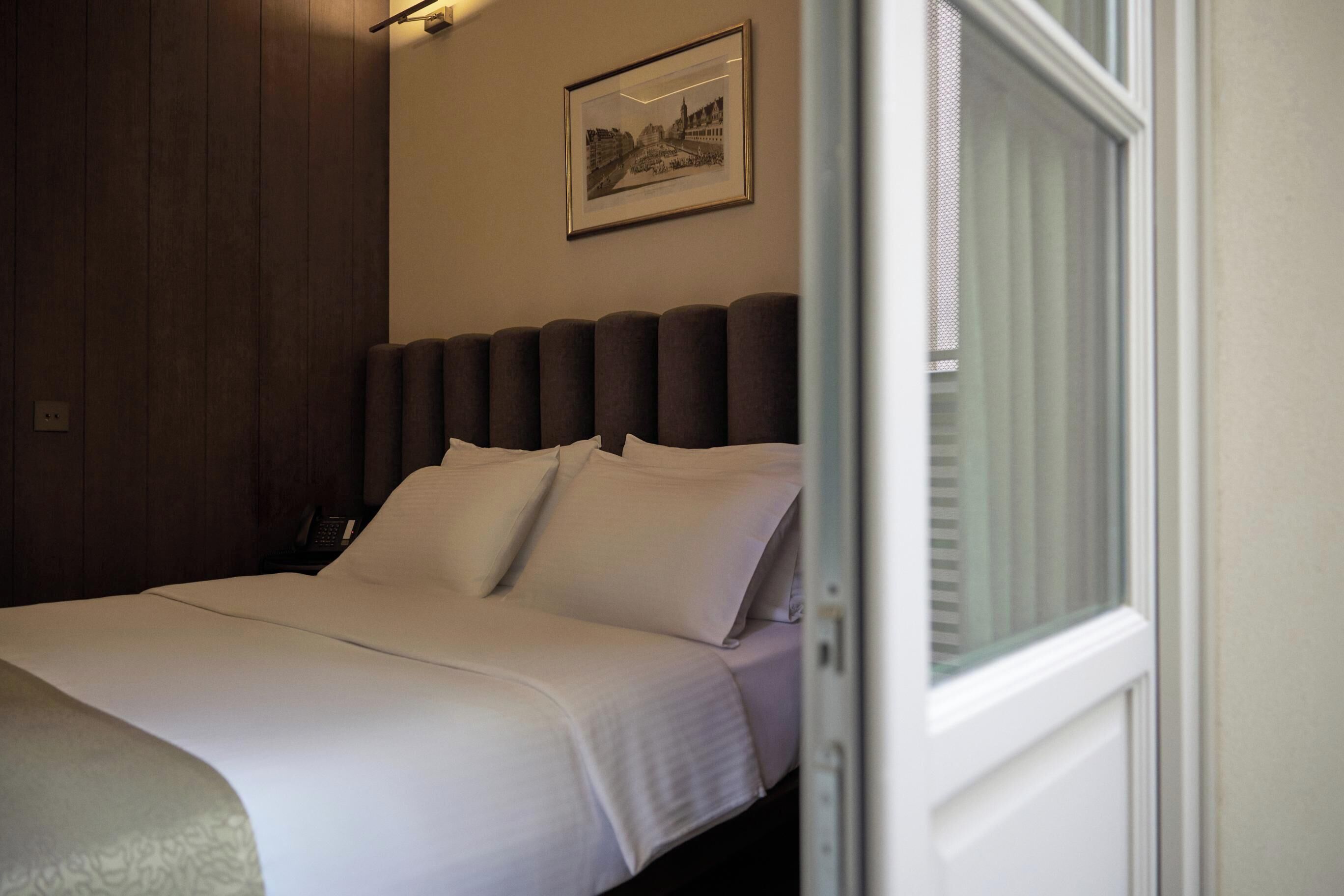 A neatly made bed with white linens and a headboard, a framed picture on the wall, and a window with blinds partially open.