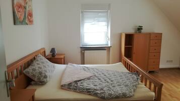 Ein Schlafzimmer mit Bett, Nachttisch, Kommode, einem Fenster mit Jalousien und einer Heizung.