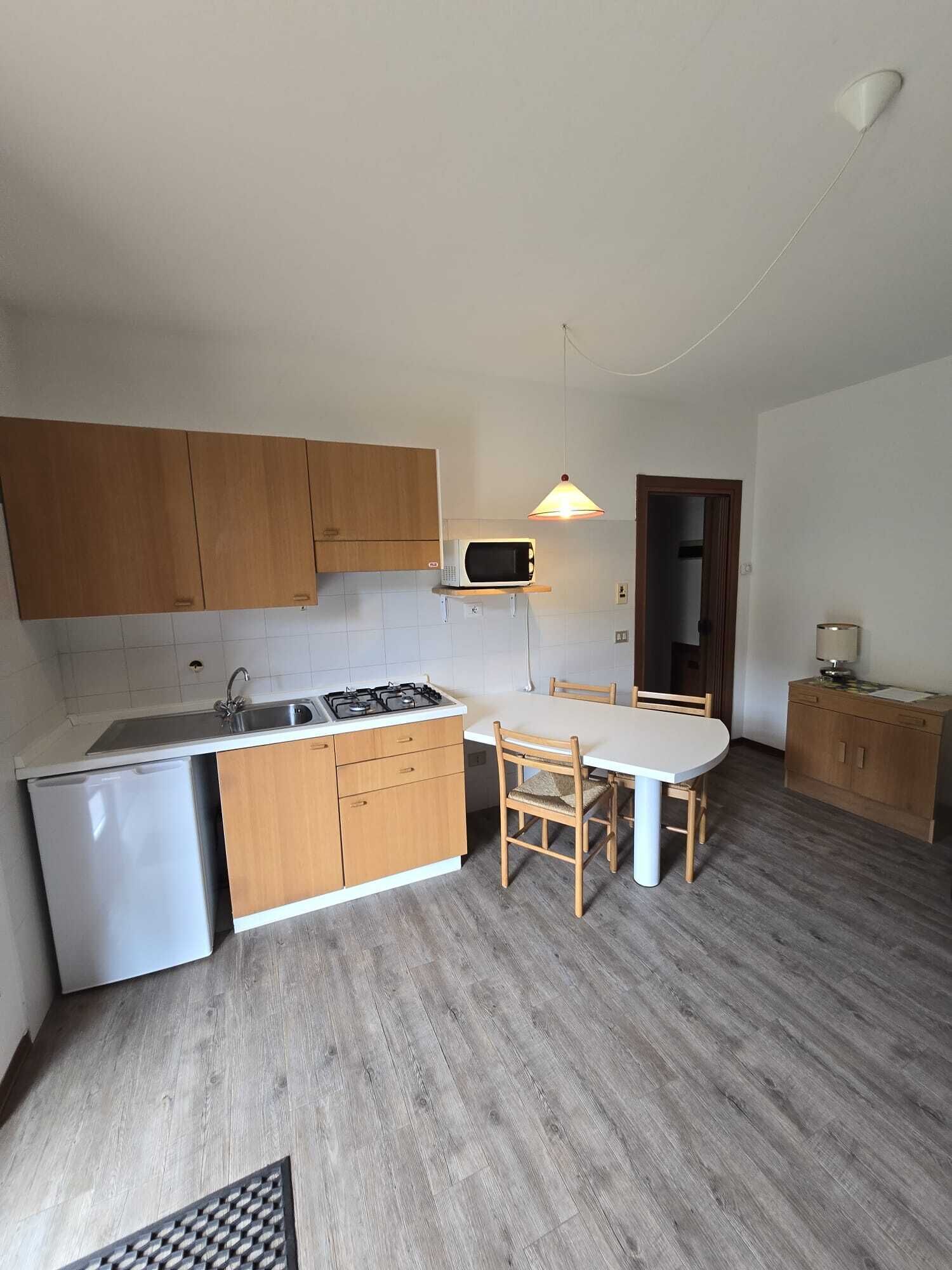 A compact kitchen with a stove, sink, and microwave, wooden cabinets, a dining table with chairs, and a hanging light fixture.
