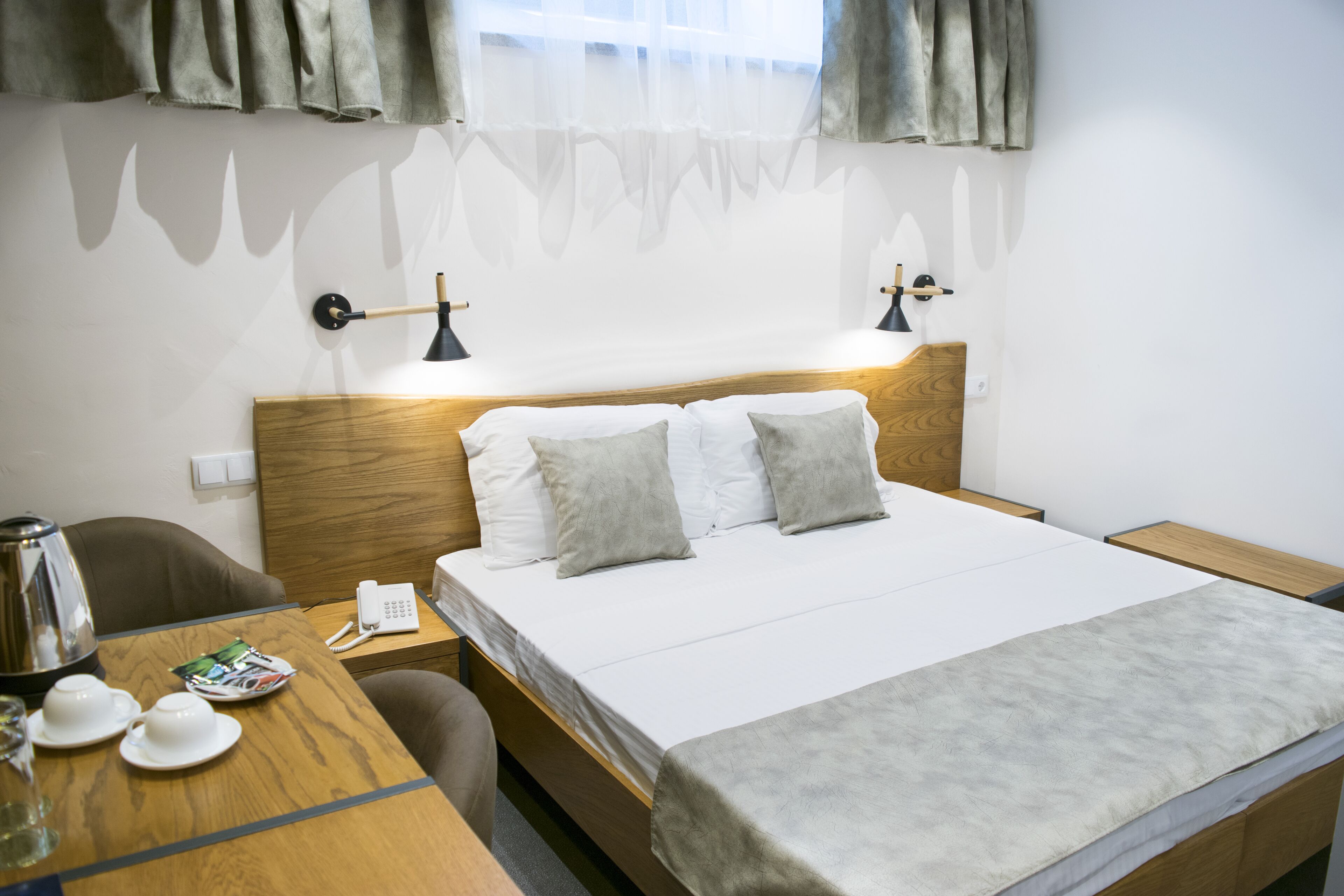 Quarto de hotel com cama de madeira, duas luminárias laterais, telefone ao lado da cama, mesa com chaleira, xícaras e uma revista.