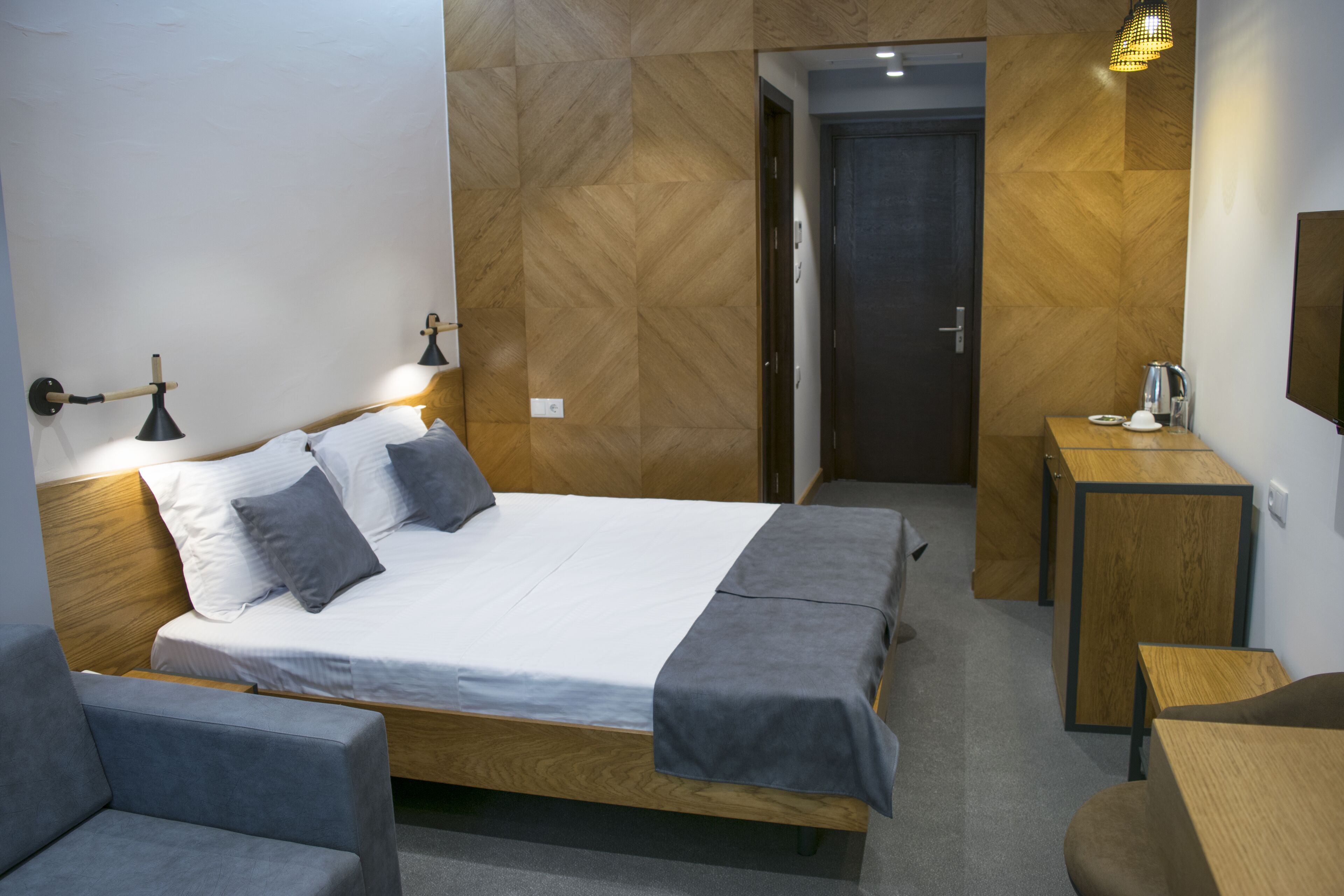 Una habitación de hotel moderna con cabecera de madera, cama con ropa de cama gris, una mesita de noche y un escritorio pequeño con una silla.
