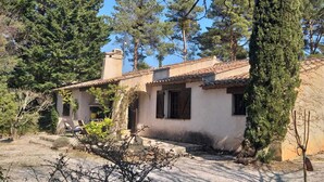 Uma casa rústica com telhado de telhas, rodeada por árvores e um caminho de acesso de cascalho.