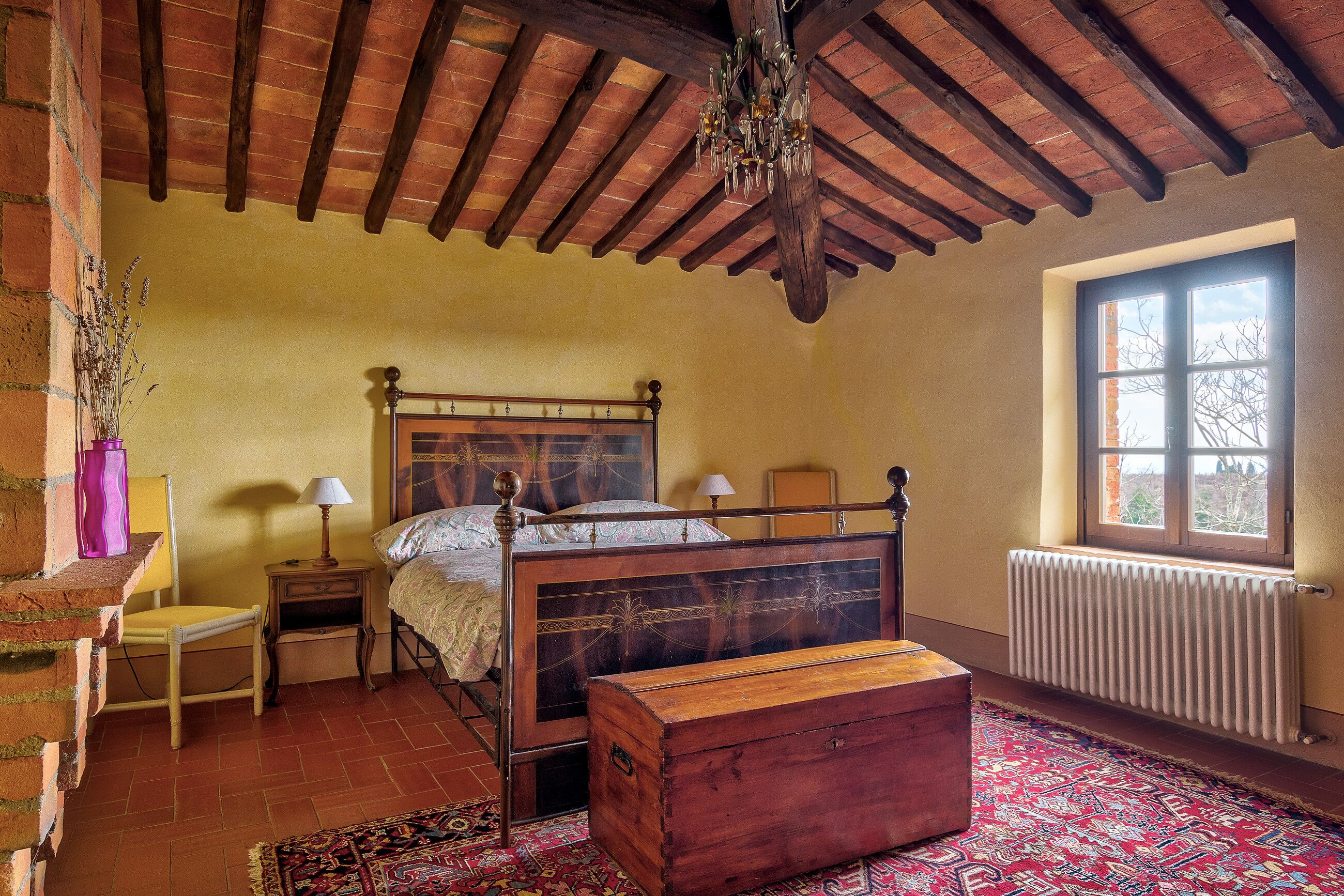 Une chambre avec un lit en bois, une commode, une fenêtre donnant sur un beau paysage, un radiateur et un tapis à motifs.