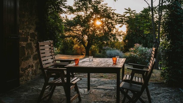 Een terras buiten met een stenen tafel en houten stoelen, omringd door groen en een stenen muur.