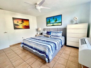 Een hotelkamer met een bed, een plafondventilator, een nachtkastje, een lamp, een dressoir en twee decoratieve wandobjecten.