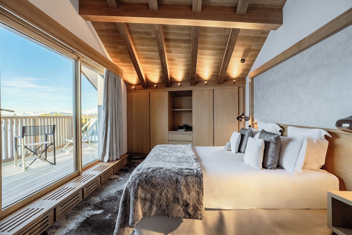 Une chambre avec une grande fenêtre, des poutres apparentes en bois au plafond, un lit avec des oreillers et un banc placé près de la fenêtre.