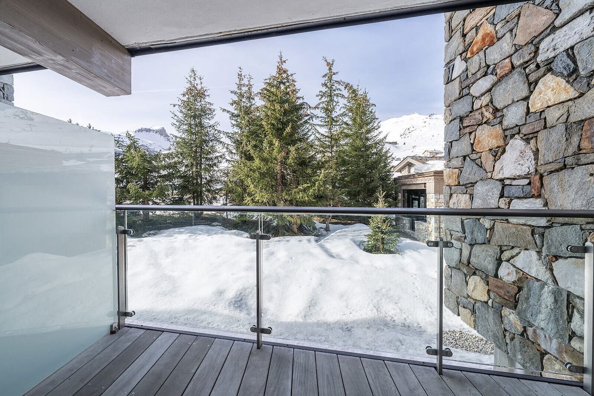 Un balcon doté d’une balustrade en verre, offrant une vue sur un paysage montagneux enneigé et un mur en pierre.