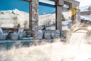Un área de spa con pared de piedra, un ventanal con vistas a una montaña nevada y una cascada.