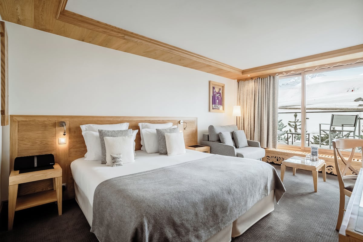 Une chambre d’hôtel avec un grand lit, une tête de lit en bois, une table de chevet avec une lampe, une chaise, une petite table et une vue sur un paysage enneigé par de grandes fenêtres.