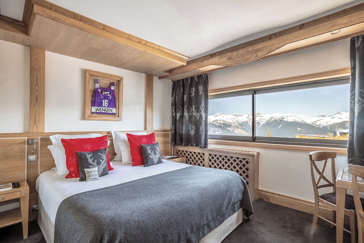 Une chambre d’hôtel avec un lit, une fenêtre donnant sur des montagnes enneigées, une tête de lit en bois et un maillot encadré accroché au mur.