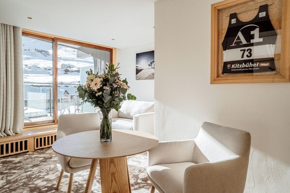 Una habitación de hotel con un ventanal grande, una mesa redonda de madera, dos sillones blancos y una camiseta enmarcada en la pared.