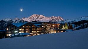 Un centro turístico en la montaña cubierto de nieve, con edificios iluminados y una luna visible en el cielo.