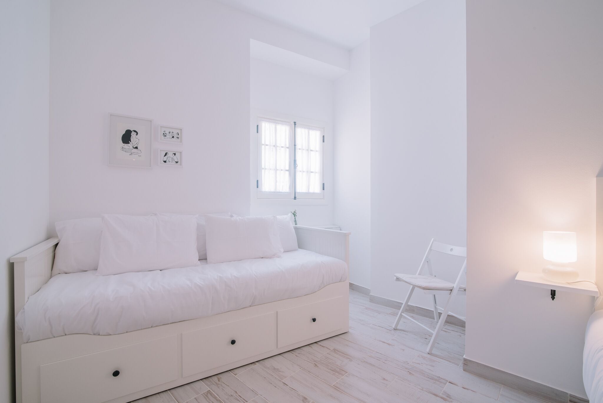 Camera da letto minimalista con un letto bianco, una scrivania e una sedia. Alle pareti ci sono quadri incorniciati e una finestra con persiane.