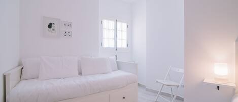 Camera da letto minimalista con un letto bianco, una scrivania e una sedia. Alle pareti ci sono quadri incorniciati e una finestra con persiane.