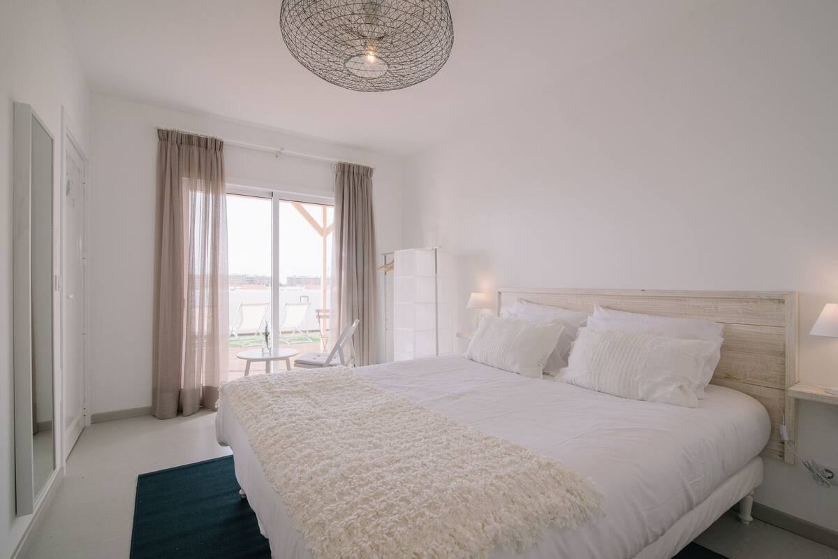 Une chambre avec un grand lit, une table de chevet avec une lampe, une fenêtre avec des rideaux et une vue sur l’extérieur.