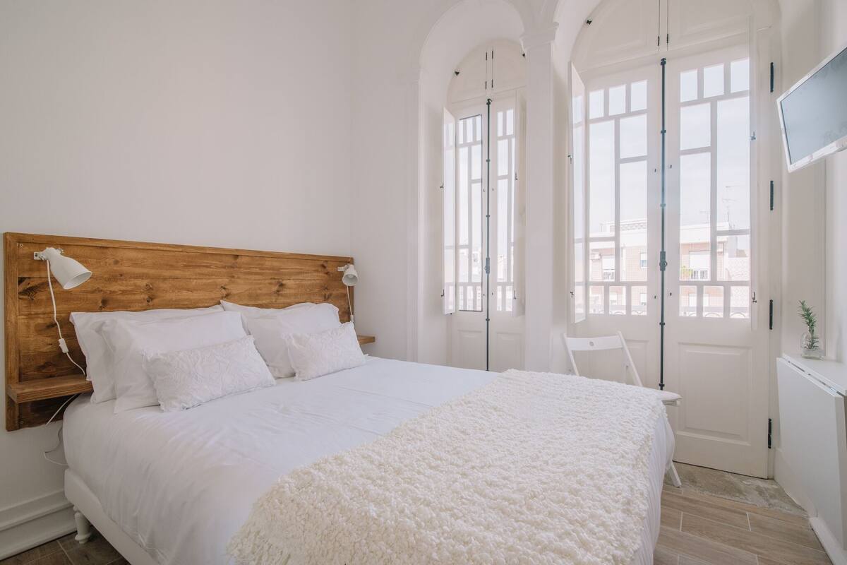 Une chambre à coucher avec une tête de lit en bois, du linge de lit blanc, un tapis blanc et un téléviseur posé sur un meuble.