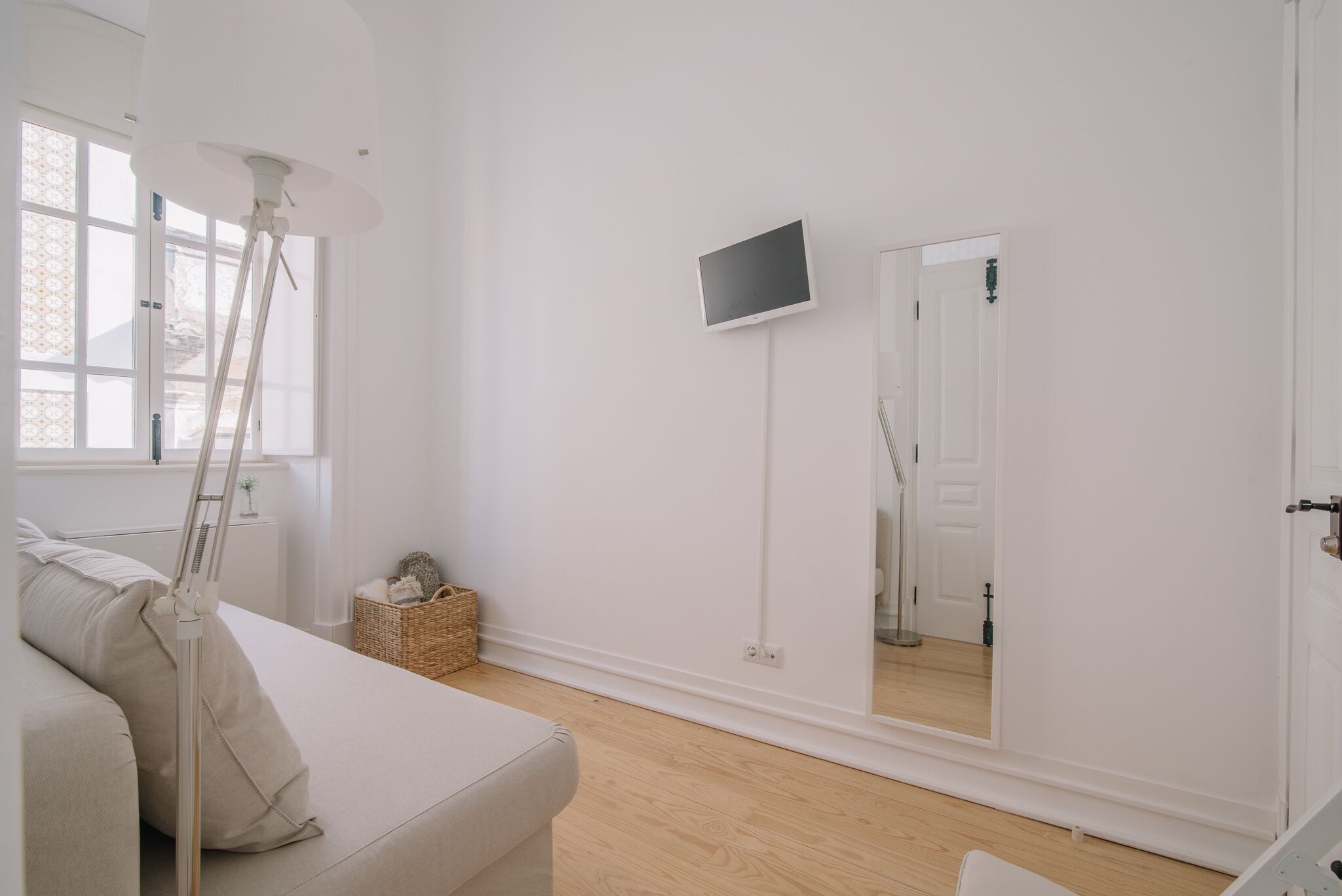 Ein schlicht eingerichteter Raum mit einem weißen Sofa, einer Stehlampe, einem an der Wand befestigten Flachbildfernseher und einem großen Spiegel, der die Tür widerspiegelt.
