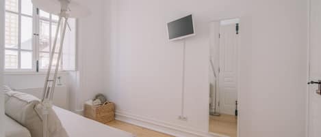 A minimalist room with a white sofa, a standing lamp, a flat-screen TV mounted on the wall, and a large mirror reflecting the door.