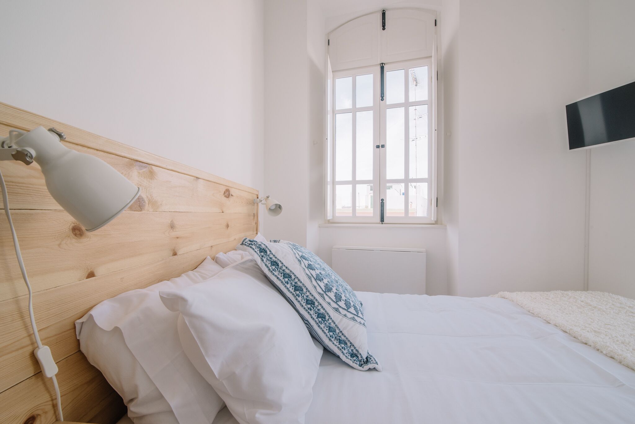 A bedroom with a wooden headboard, a bedside lamp, a window, and a flat-screen TV mounted on the wall.