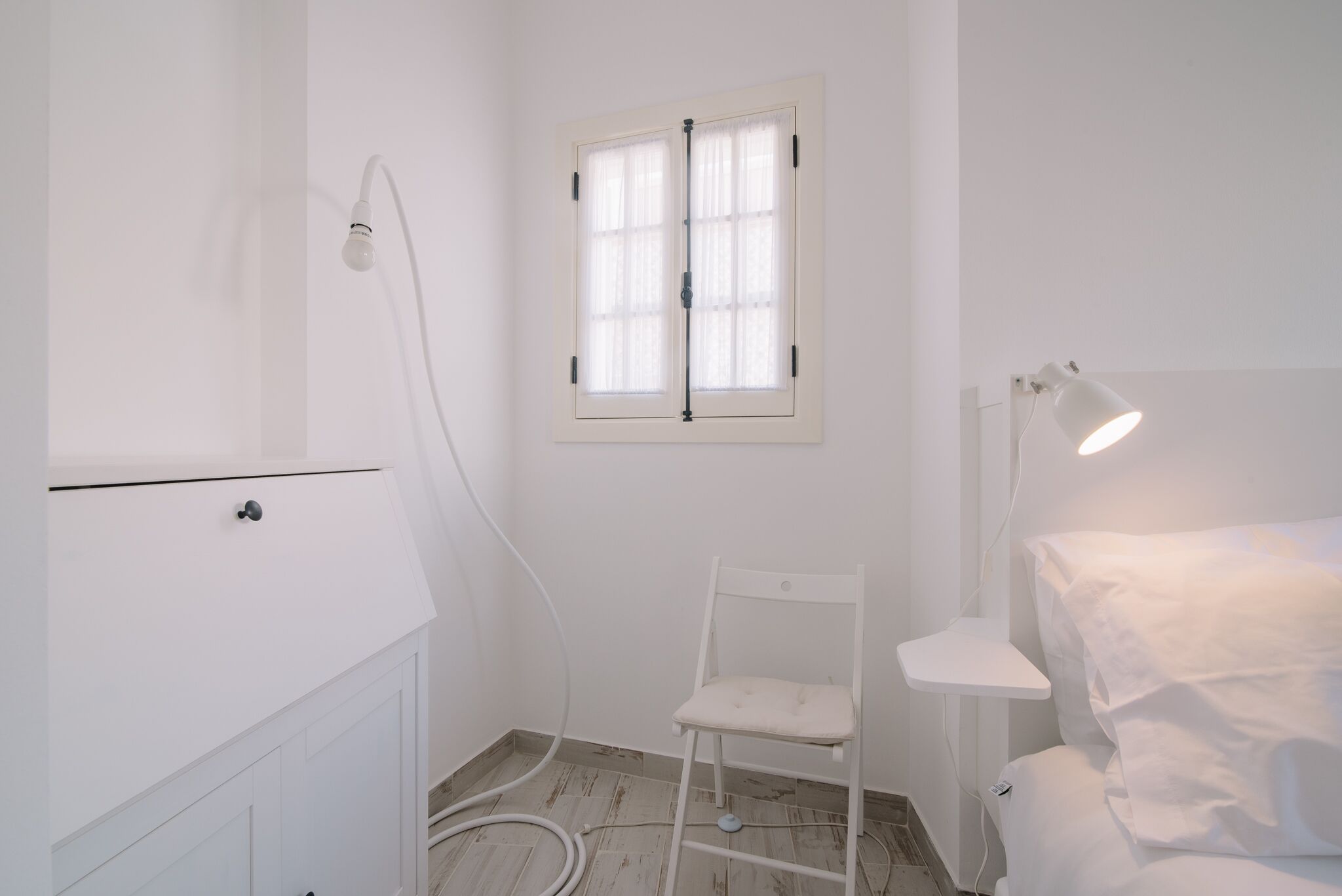 A white room with a window, a chair, a bedside table, and a lamp.