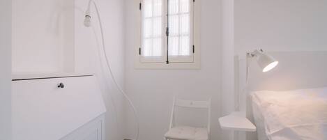 A white room with a window, a chair, a bedside table, and a lamp.