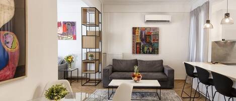 A modern living room with a grey sofa, a glass coffee table, and a dining area with a white table and black chairs.