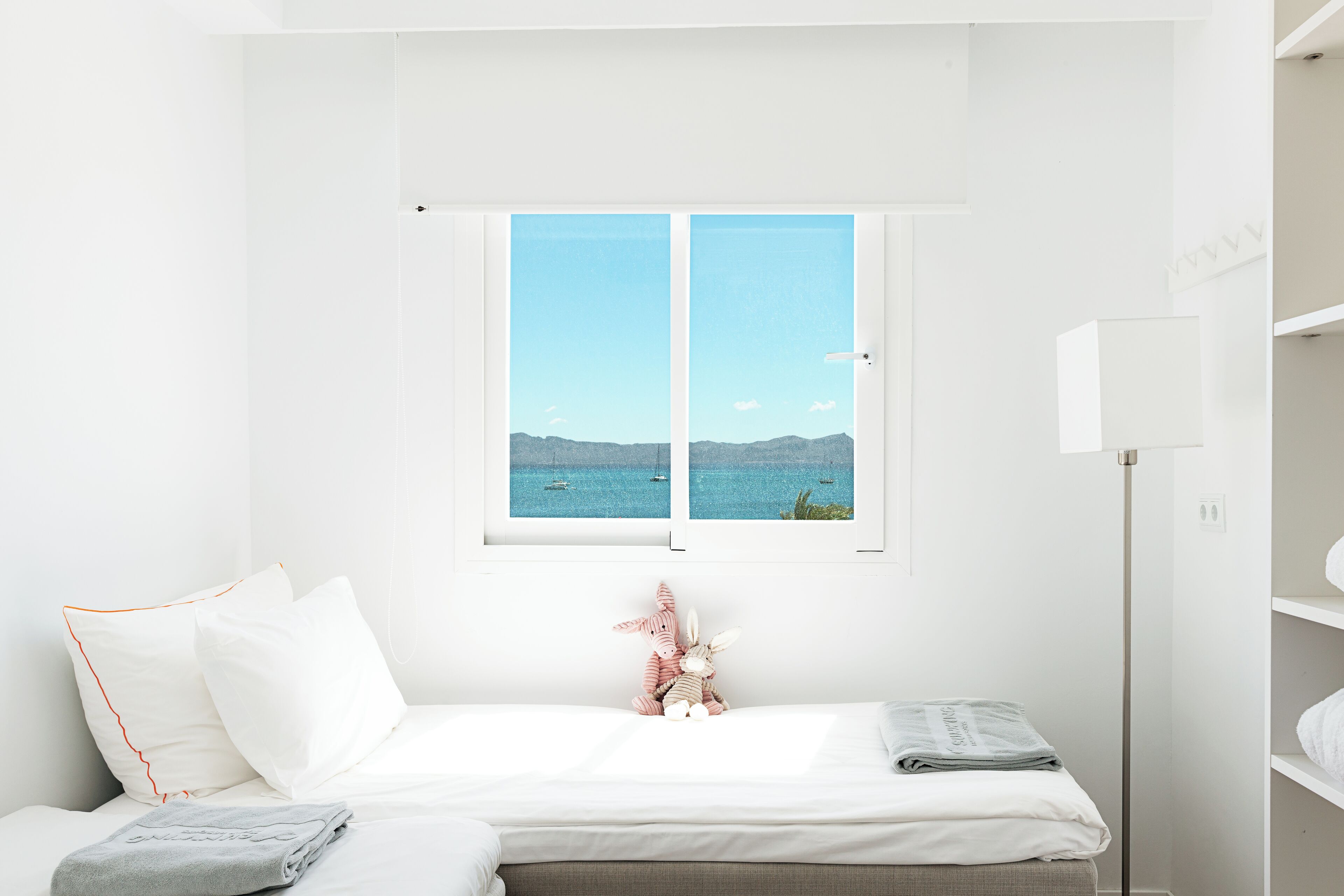 Una habitación minimalista con una cama, un ventanal con vistas al mar y un estante pequeño.