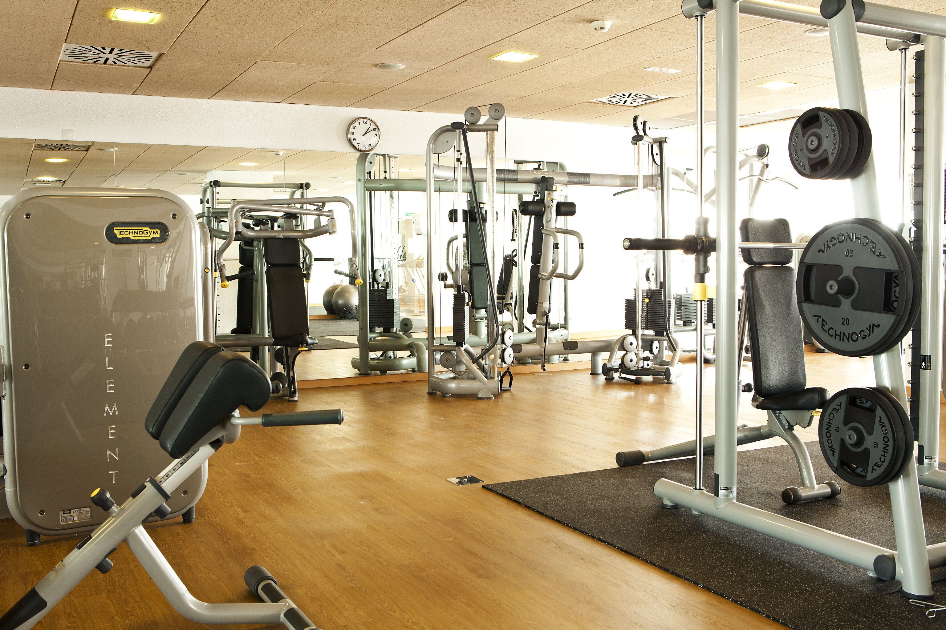 A gym with various exercise machines, a clock on the wall, and a wooden floor.