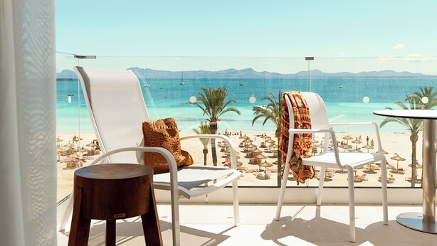 Un balcón con dos sillas blancas, una mesa redonda pequeña y vistas a una playa con sombrillas y palmeras.